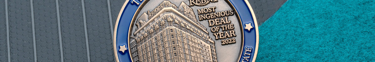 Close crop of a blue-rimmed 3D coin showing the sculpted building relief and the REBNY Most Ingenious Deal of the Year 2022 award text