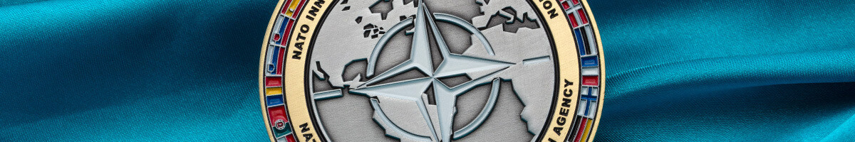 Close-up of the silver NATO compass star raised over a world map, framed by a gold ring of member-nation flags